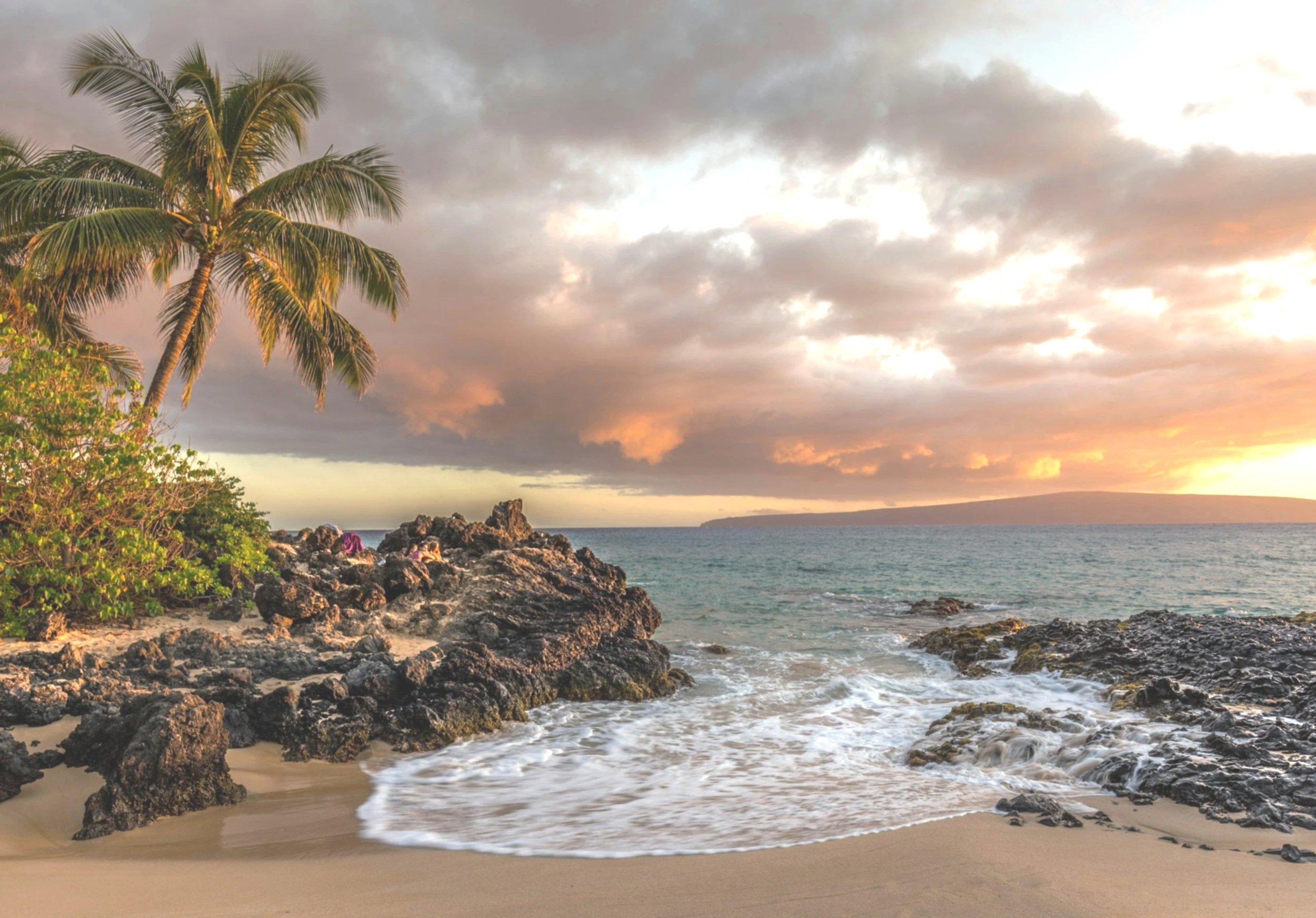 Tropical beach at sunset with palm trees, rocky shoreline, sandy beach, ocean waves, and a colorful sky with clouds.