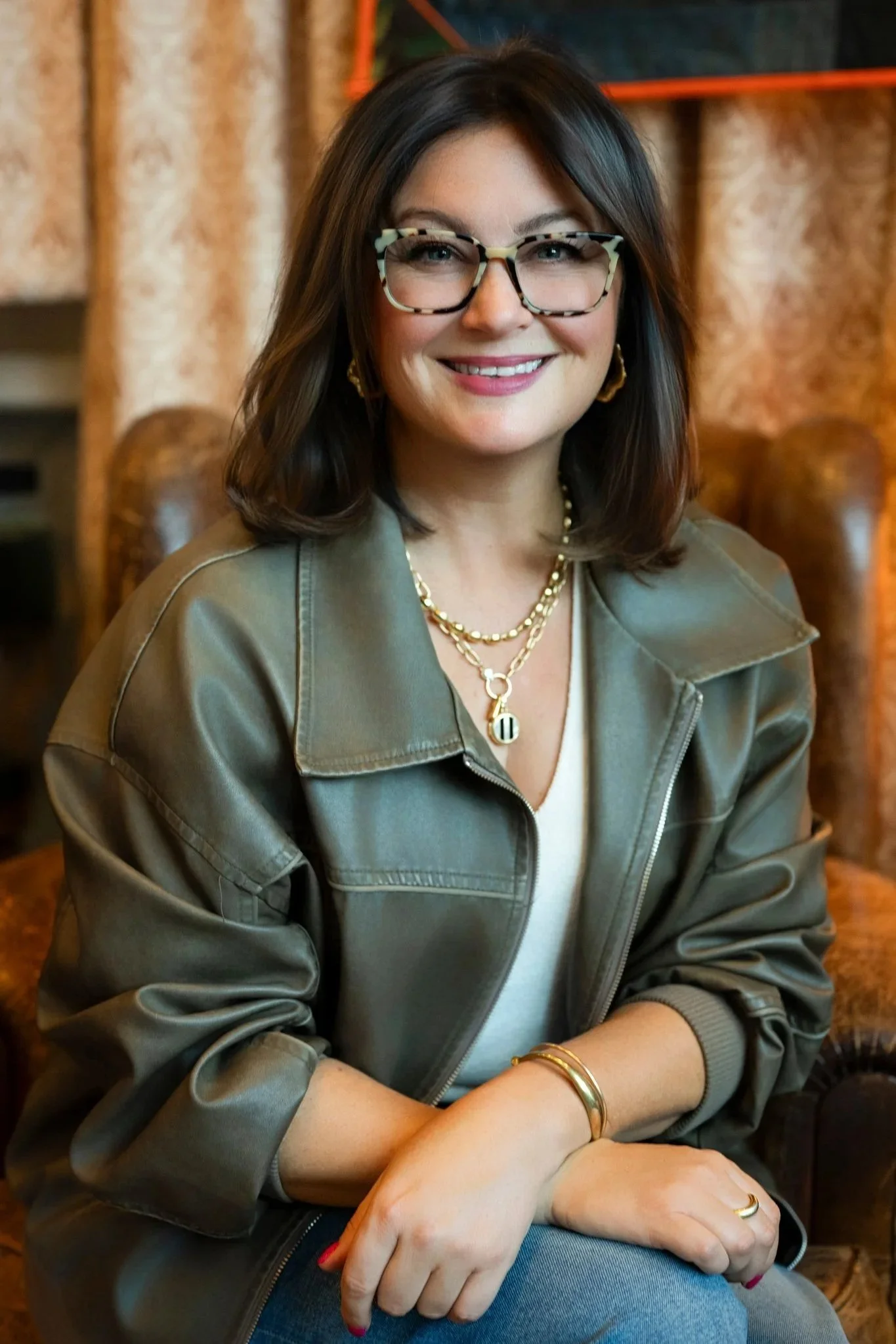 A woman with short dark hair, large tortoise shell glasses, and gold jewelry, smiling while seated indoors with wooden furniture and patterned curtains in the background.