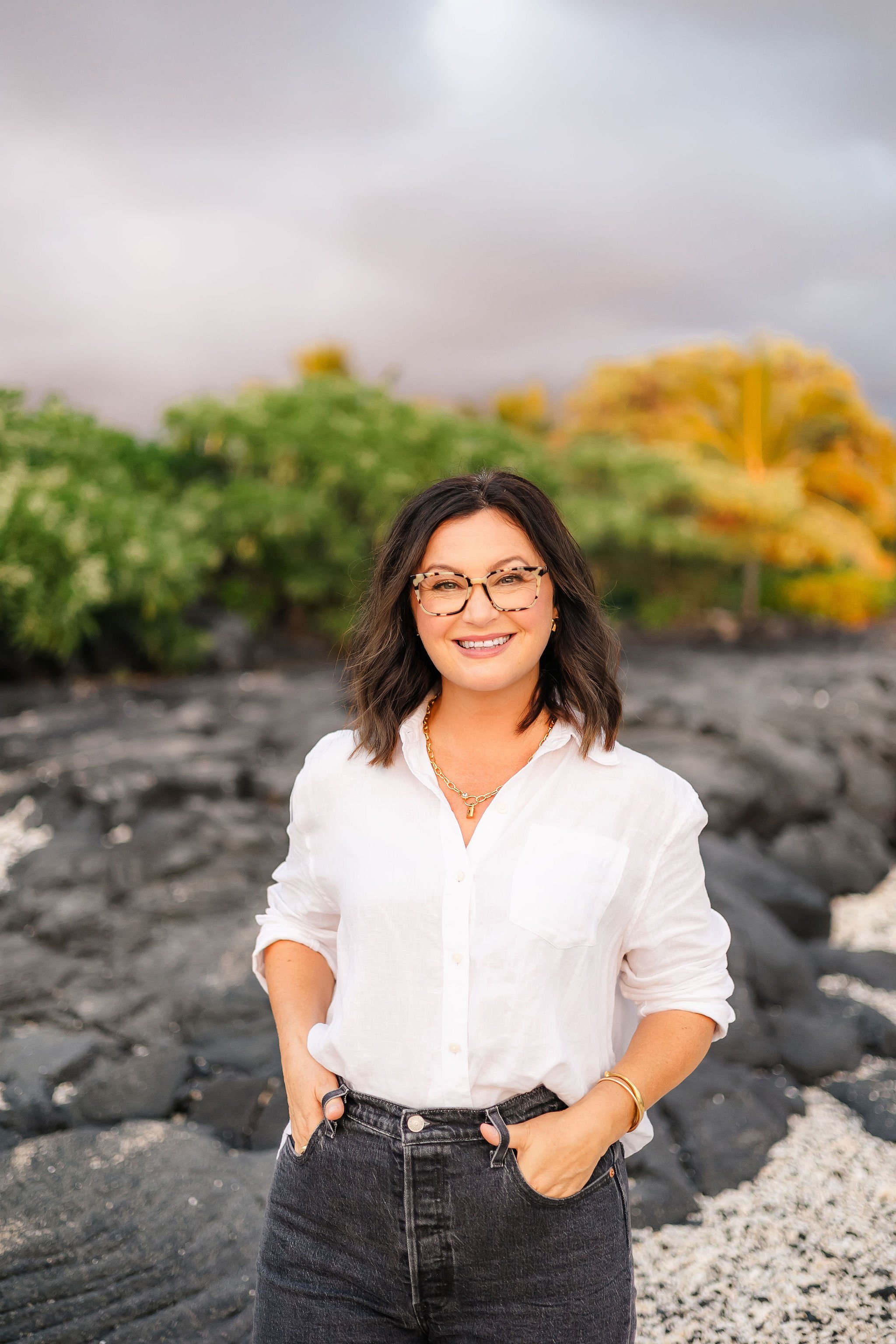 A woman with dark hair and glasses smiling outdoors, wearing a white shirt and black jeans, standing on a rocky landscape with trees in the background.