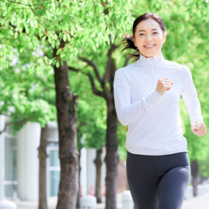 A woman jogging outdoors in a park with green trees, wearing a white long-sleeve shirt and black leggings, smiling.