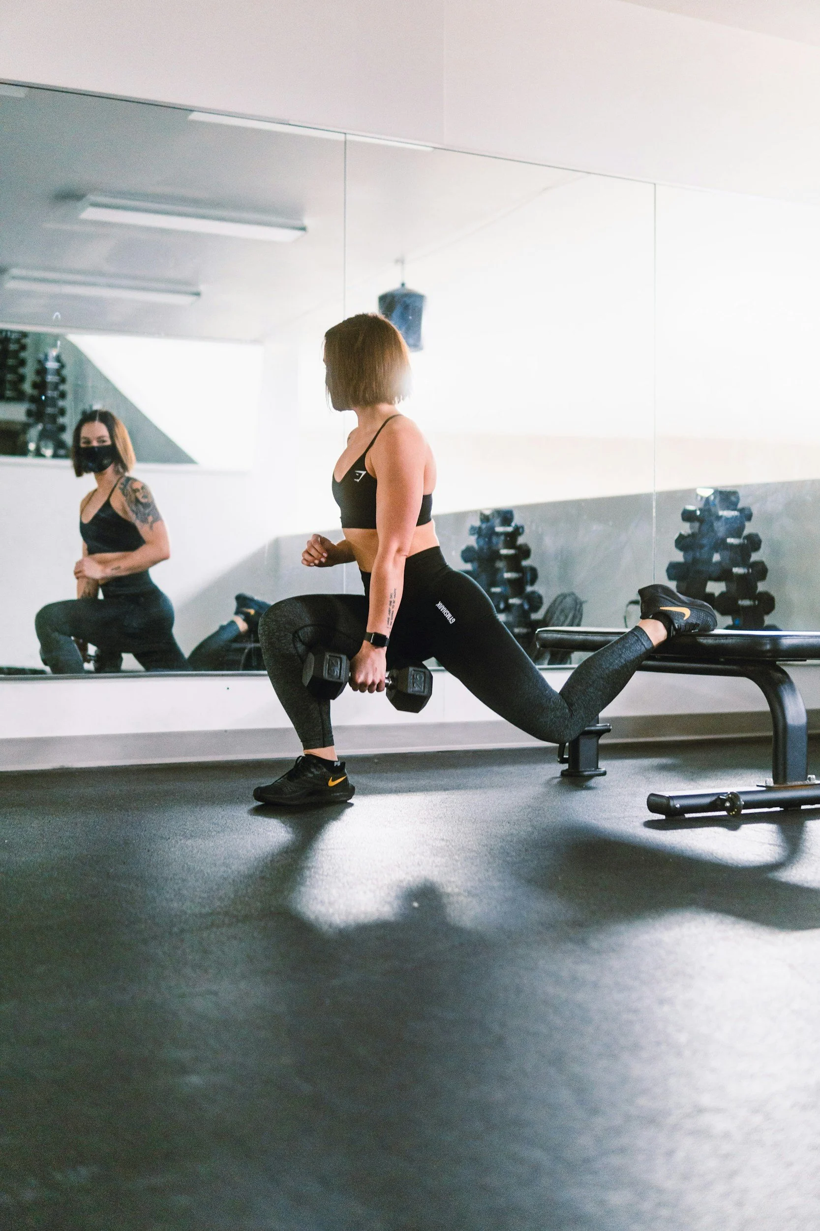 A woman in workout attire performs a lunge exercise while holding dumbbells in a gym, with her reflection visible in a mirror behind her.