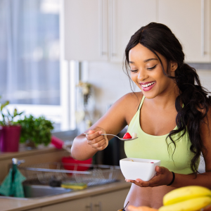 A woman with long black hair in a ponytail, wearing a yellow tank top, smiling while eating cereal from a white bowl in a bright kitchen.