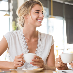 A smiling woman with blonde hair holding a white mug while sitting at a table in a well-lit cafe.