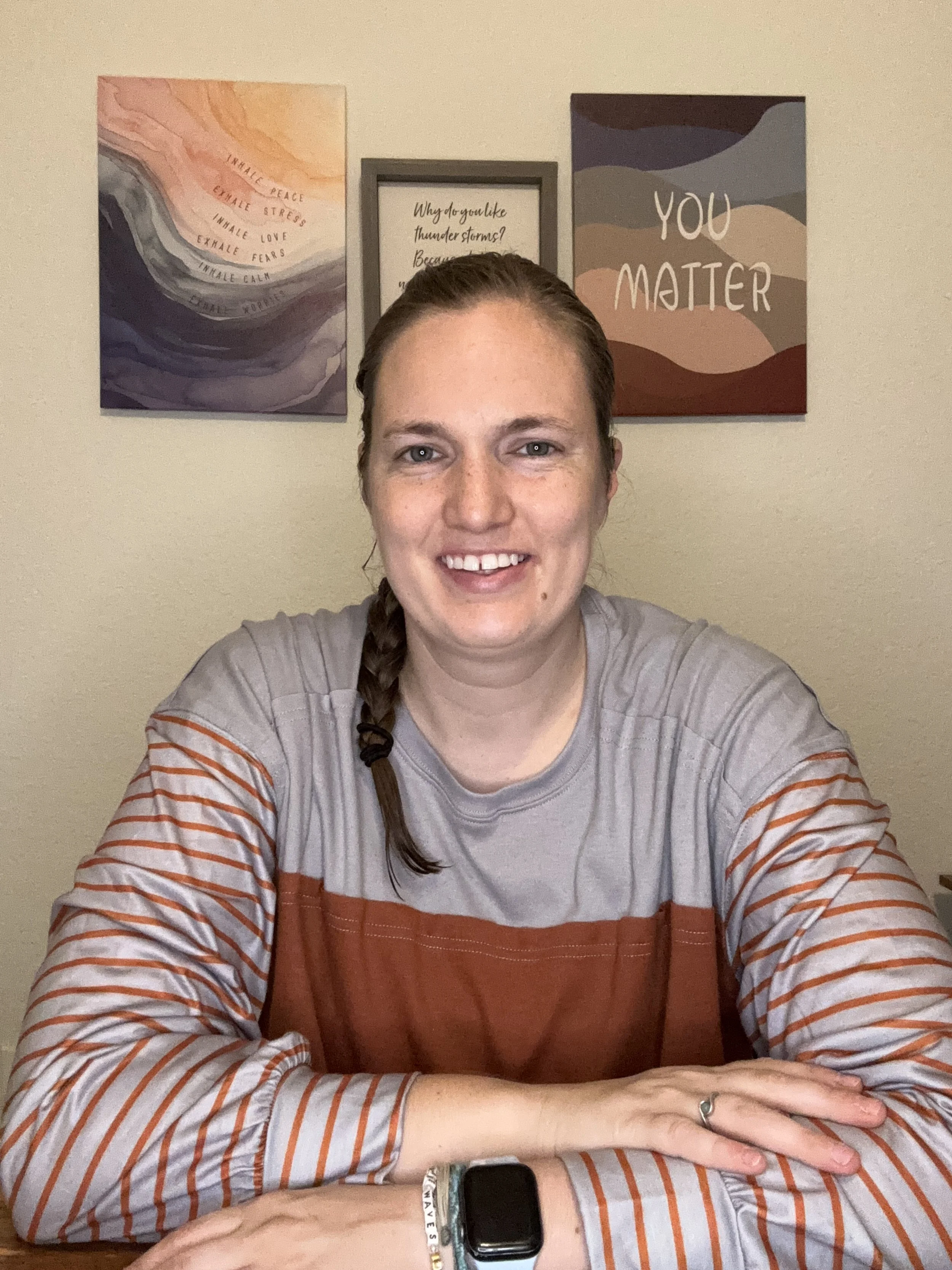 A woman with a braid smiling, sitting at a table with her hands resting on it, wearing a striped shirt and a watch, with three motivational posters on the wall behind her.