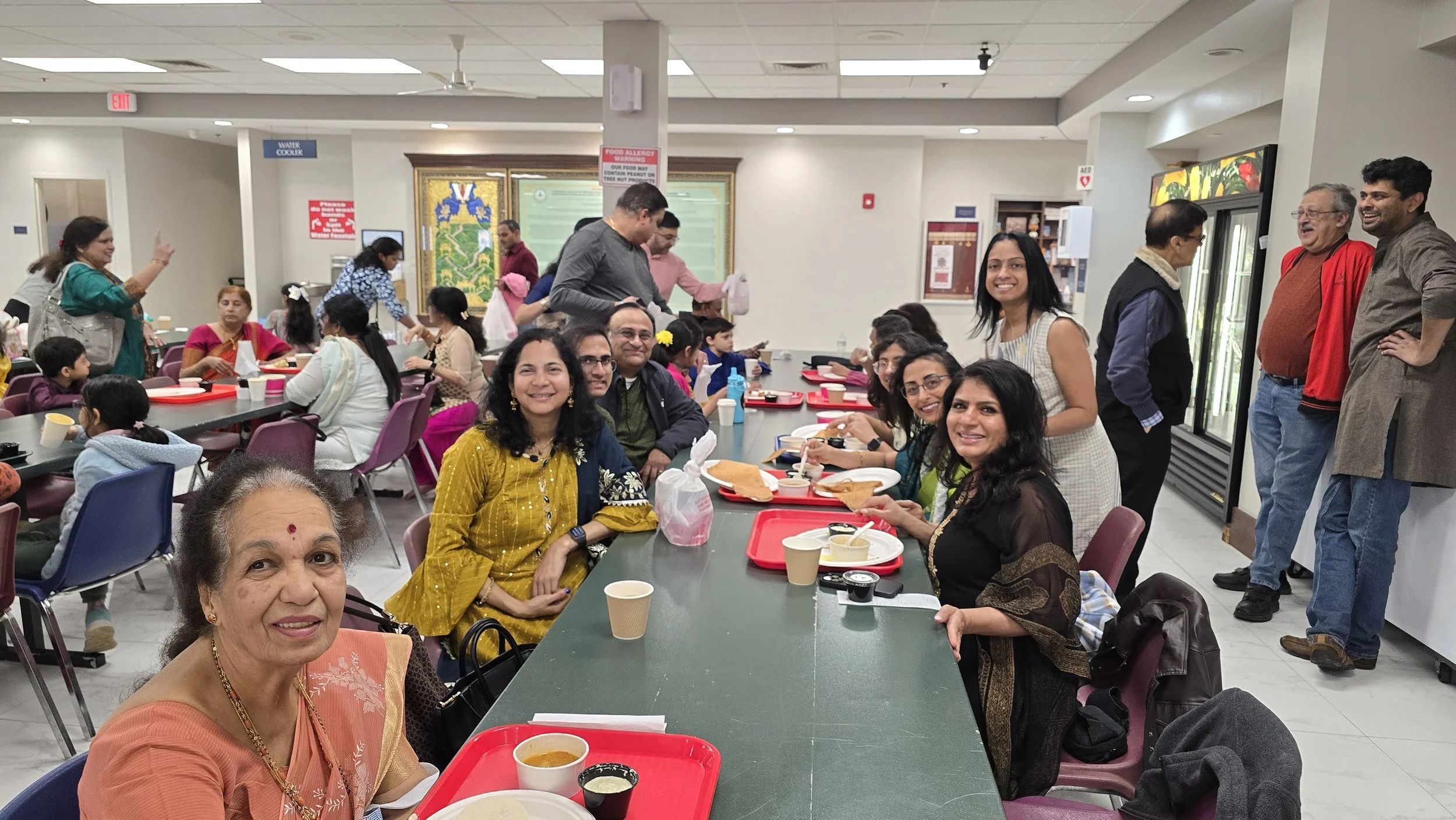 People playing carrom in an indoor sports hall, with others in the background participating in table tennis. Some individuals are seated, watching, or standing, observing the games.