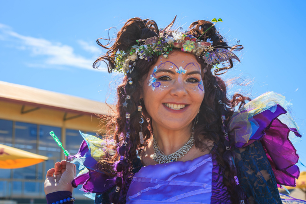 Woman dressed in a colorful fairy costume with face paint and a flower crown, smiling outdoors on a sunny day.