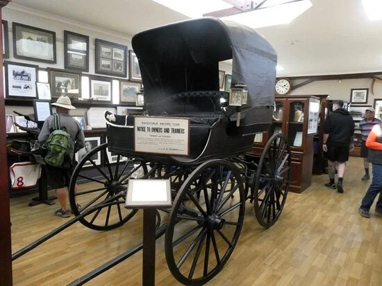 A vintage black horse-drawn carriage displayed indoors at a museum, with people viewing exhibits around it.