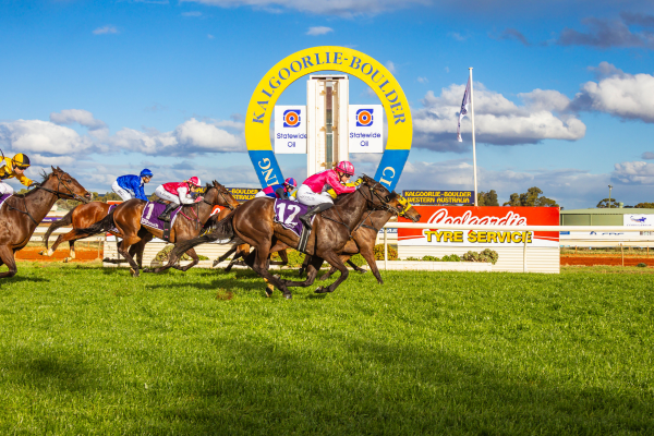 Horse race at a track with jockeys in colorful uniforms, with a large round sign in the background that reads Kalgoorlie-Boulder.