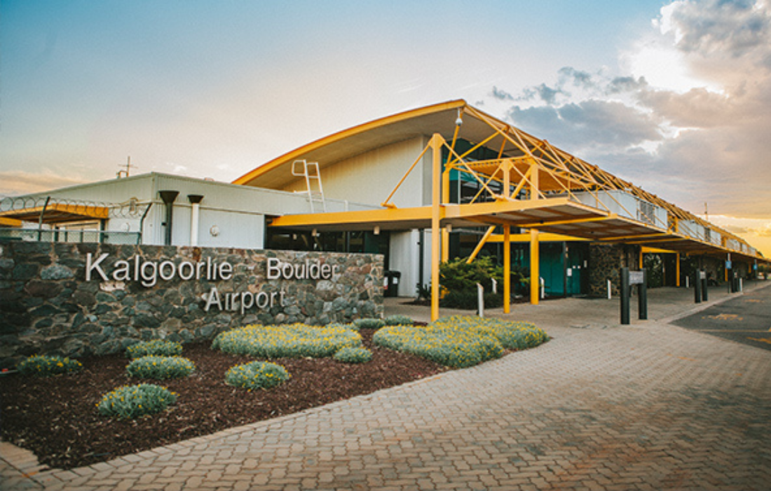 Exterior view of Kalgoorlie Boulder Airport with a stone sign and flower bed in the foreground, and a modern building structure with yellow accents under a partly cloudy sky.