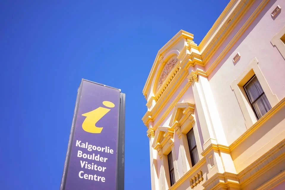 Sign for the Kalgoorlie Boulder Visitor Centre next to a historic building with ornate yellow and white architectural details under a clear blue sky.