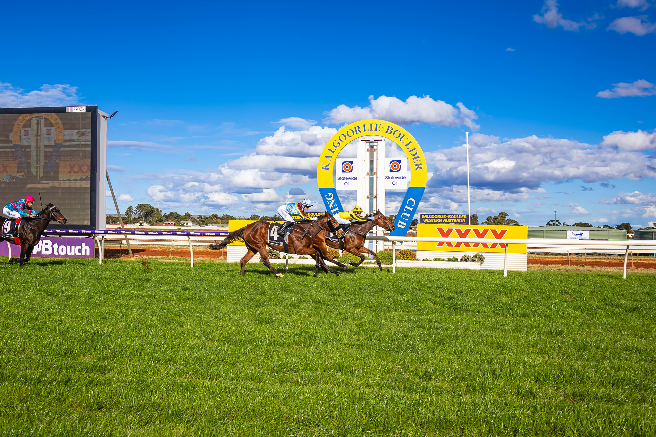 Horse race at Kalgoorlie-Boulder Racecourse in Western Australia with four horses running on the track under a partly cloudy blue sky.