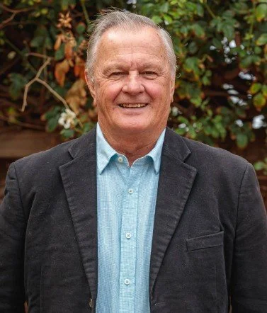 A smiling older man in a blue button-up shirt and dark blazer standing outdoors with green foliage in the background.