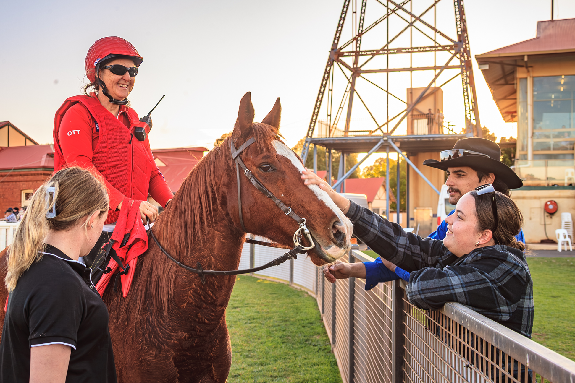 Three people, including a woman in a red jacket and riding gear, are interacting with a brown horse at a fairground during the sunset. The woman is sitting on the horse, and two people on the other side of the fence are petting and reaching out to the horse. There are buildings and a tower structure in the background.