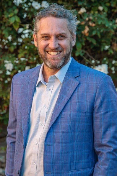 A smiling man with gray curly hair and a beard, wearing a blue plaid blazer and a light blue collared shirt, standing outdoors in front of greenery.