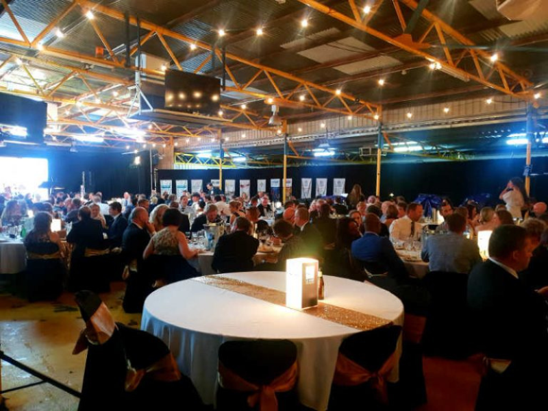 A large indoor banquet hall filled with many seated guests at round tables, decorated with white tablecloths and centerpieces, under a ceiling with orange beams and string lighting, attending a formal event or conference.