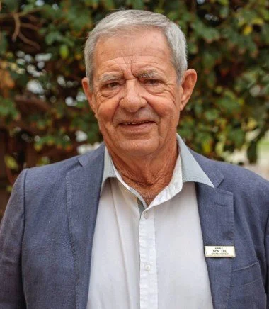 A senior man with gray hair wearing a navy blazer and white shirt standing outdoors in front of greenery.