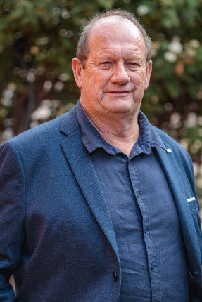 A middle-aged man in a blue blazer and shirt standing outdoors with greenery in the background.