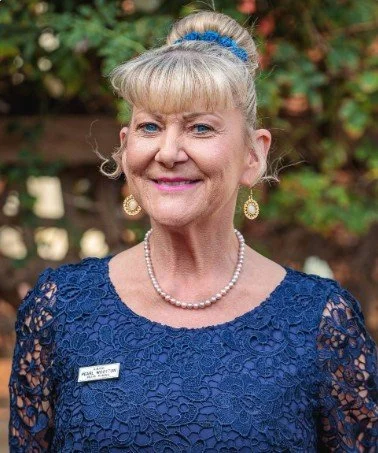 An older woman with blonde hair in a bun, wearing a blue lace dress, pearl necklace, earrings, and a blue hair accessory, smiling outdoors with greenery in the background.