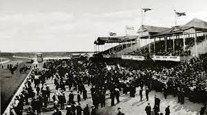 Historical black-and-white photo of a large crowd gathered at a beach or boardwalk, with old-style buildings and flags visible in the background.