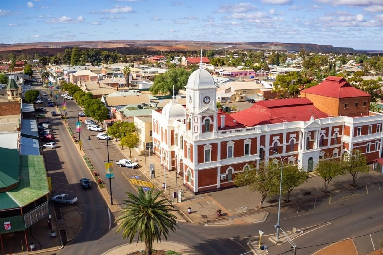 Aerial view of a city street with a historic red brick building featuring a white clock tower, lined with trees and parked cars, with colorful banners on lamp posts and a backdrop of desert hills under a blue sky.