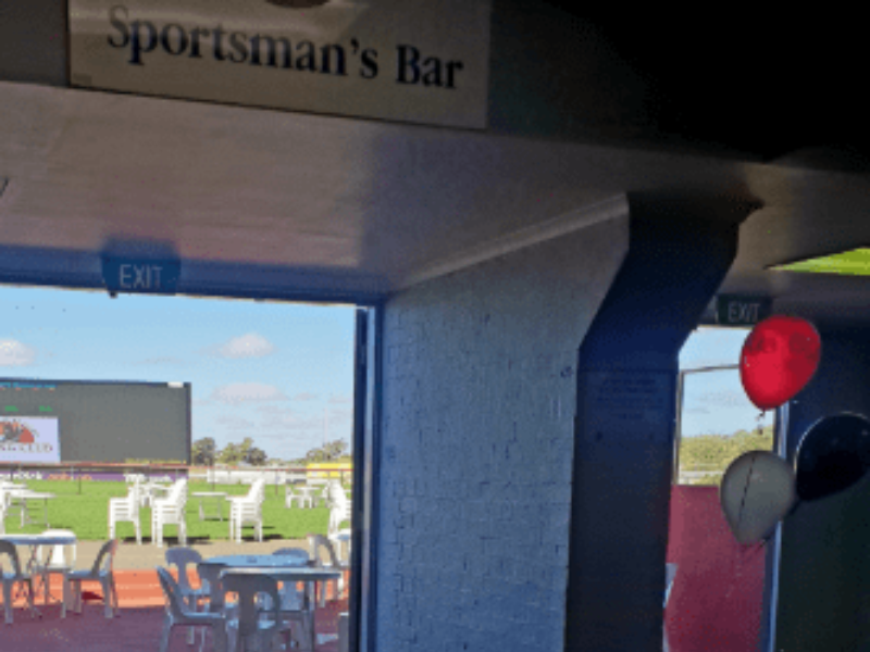 An outdoor sports bar with white furniture and a grassy area visible through the open entrance, with a blue sky and clouds.
