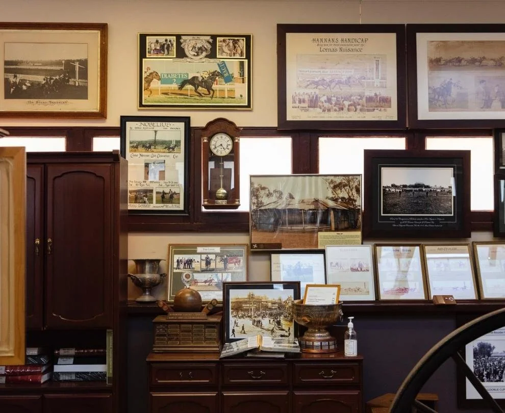 Collection of framed vintage photographs and posters on a wall and shelf in a room, with wooden cabinets and a clock.
