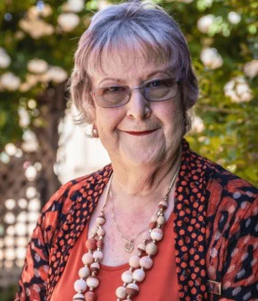 An elderly woman with short gray hair, glasses, and a warm smile, standing outdoors with greenery in the background. She is wearing a patterned jacket, a coral top, and a pearl necklace.