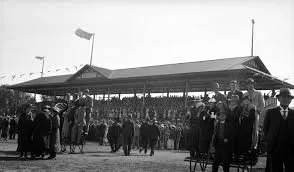 A black-and-white photo of a large crowd of people gathered at a race track, with a grandstand in the background.