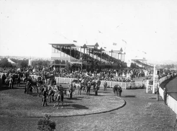 Historical black and white photo of a horse racing track with spectators' stands in the background and horses with jockeys preparing for a race on the track.