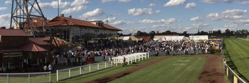A large crowd of people gathered at a race track, with a building and a grandstand in the background, under a partly cloudy sky.