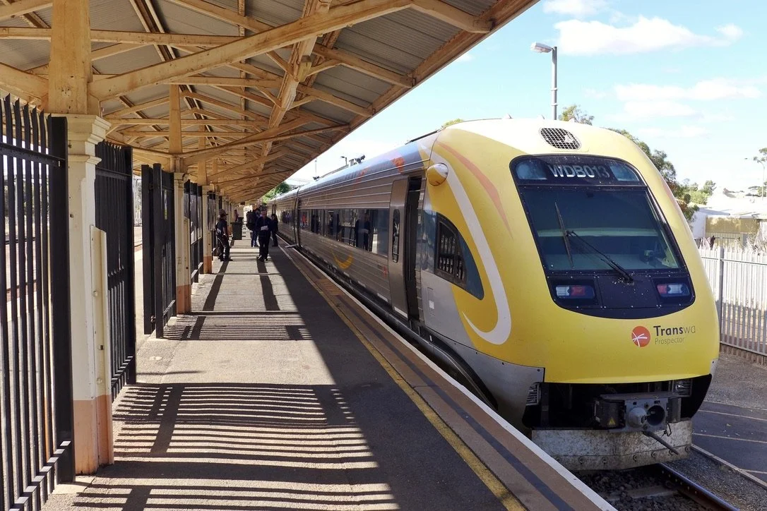 A modern yellow and gray train at a station platform with a few passengers waiting and walking.