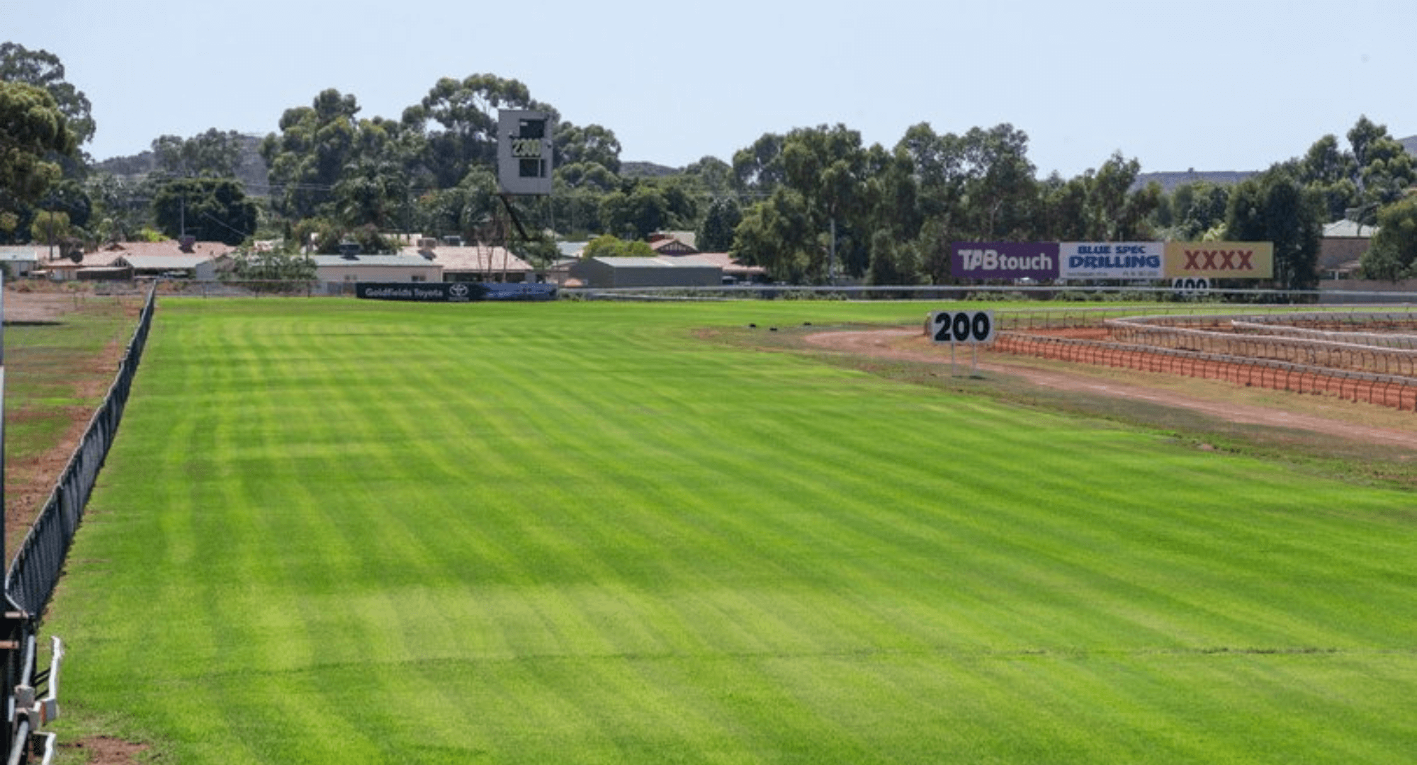 A green grassy field with a black fence on the left, dirt track on the right, and advertising billboards in the background. There is a digital scoreboard and trees beyond the field.