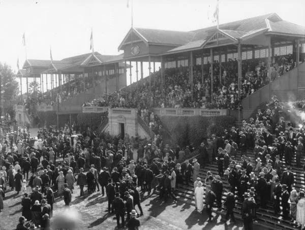 Black and white photo of a large crowd gathering outside a grand train station with a two-story covered platform, stairs leading up to the station, and many people dressed in early 20th-century clothing.