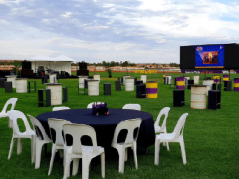 Outdoor event setup with round table and chairs, barrels and black boxes arranged on a grassy field with a stage and large screen in the background.