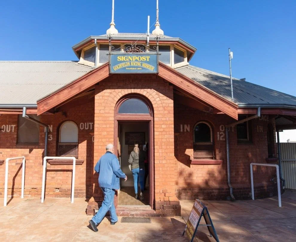 Brick building with a signpost for the Goldfields Racing Museum. Two people are walking into the museum on a sunny day.