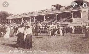 An old black-and-white photo showing a crowded outdoor event with people dressed in vintage clothing, possibly from the early 20th century, gathered on a grassy field in front of a large wooden building.