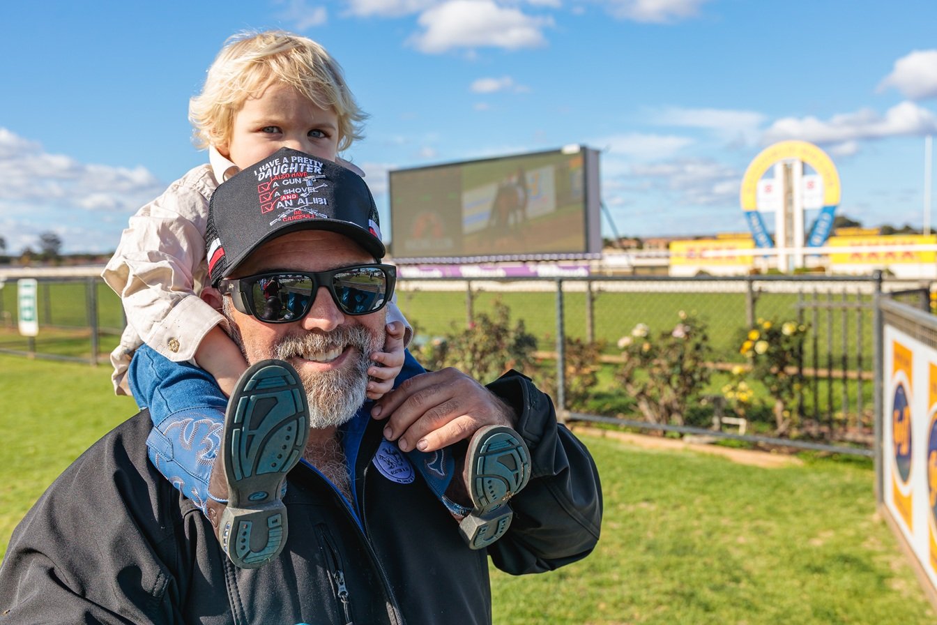 A man with a beard and sunglasses smiling with a young girl sitting on his shoulders at an amusement park or fair. The girl is holding onto his head. In the background, there is a racetrack, a large digital screen, and a colorful ride structure under a partly cloudy sky.