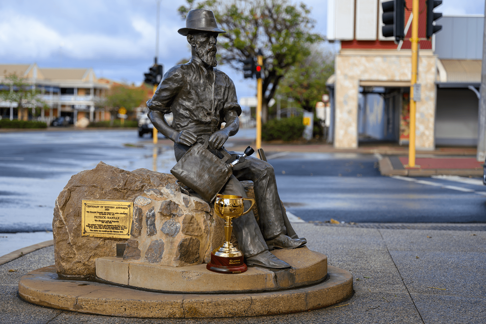 Bronze statue of a man sitting on a rock with a gold trophy placed in front of it on a city sidewalk, with buildings, trees, and traffic signals in the background.