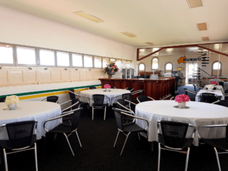 A banquet hall with decorated round tables, pink and white floral centerpieces, and a wooden bar counter in the background.