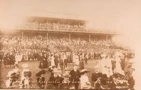 A historical photograph of a large crowd gathered at a racetrack, with a grandstand filled with spectators in the background.