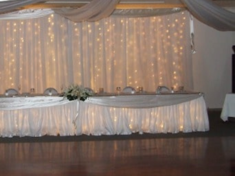 A decorated banquet table set for an event, with a white draped backdrop illuminated by string lights and the table covered with a white cloth, decorated with a floral centerpiece.
