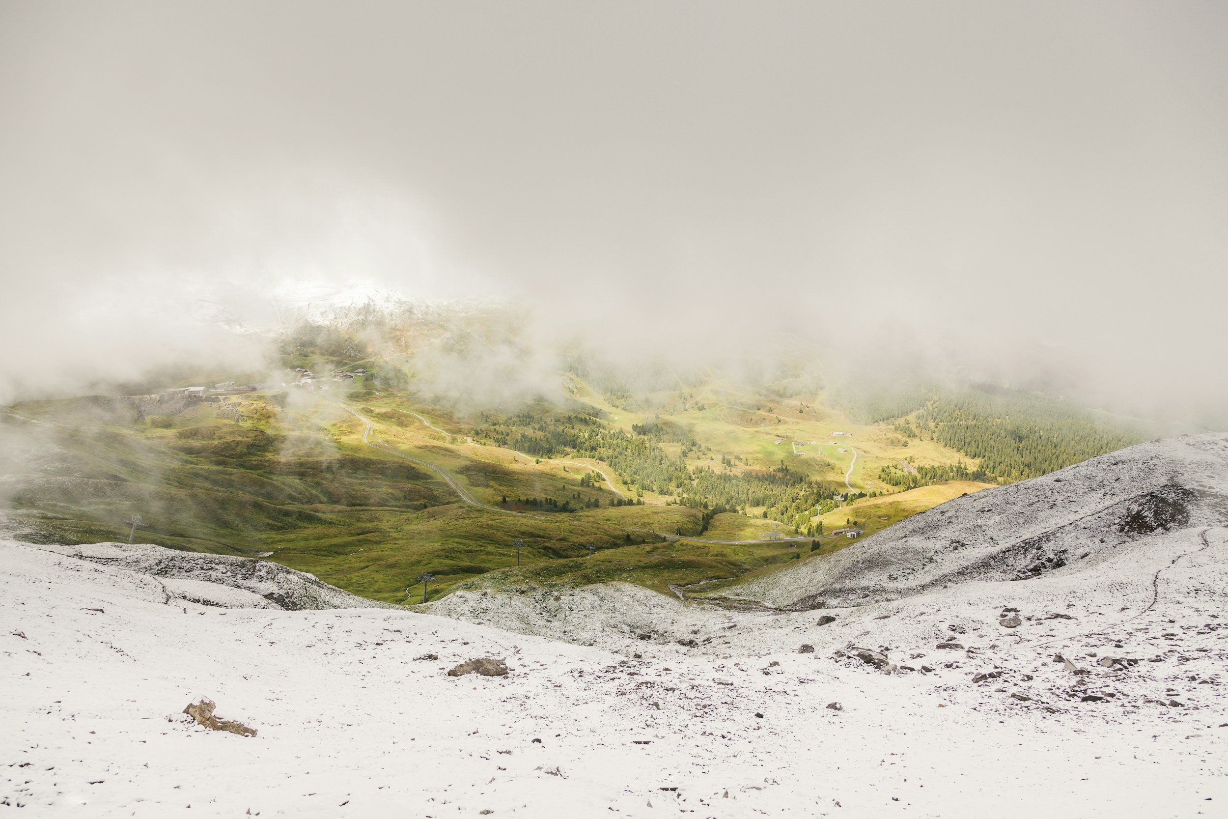 View from a snowy mountain overlooking green hills and winding roads, with clouds hanging low.