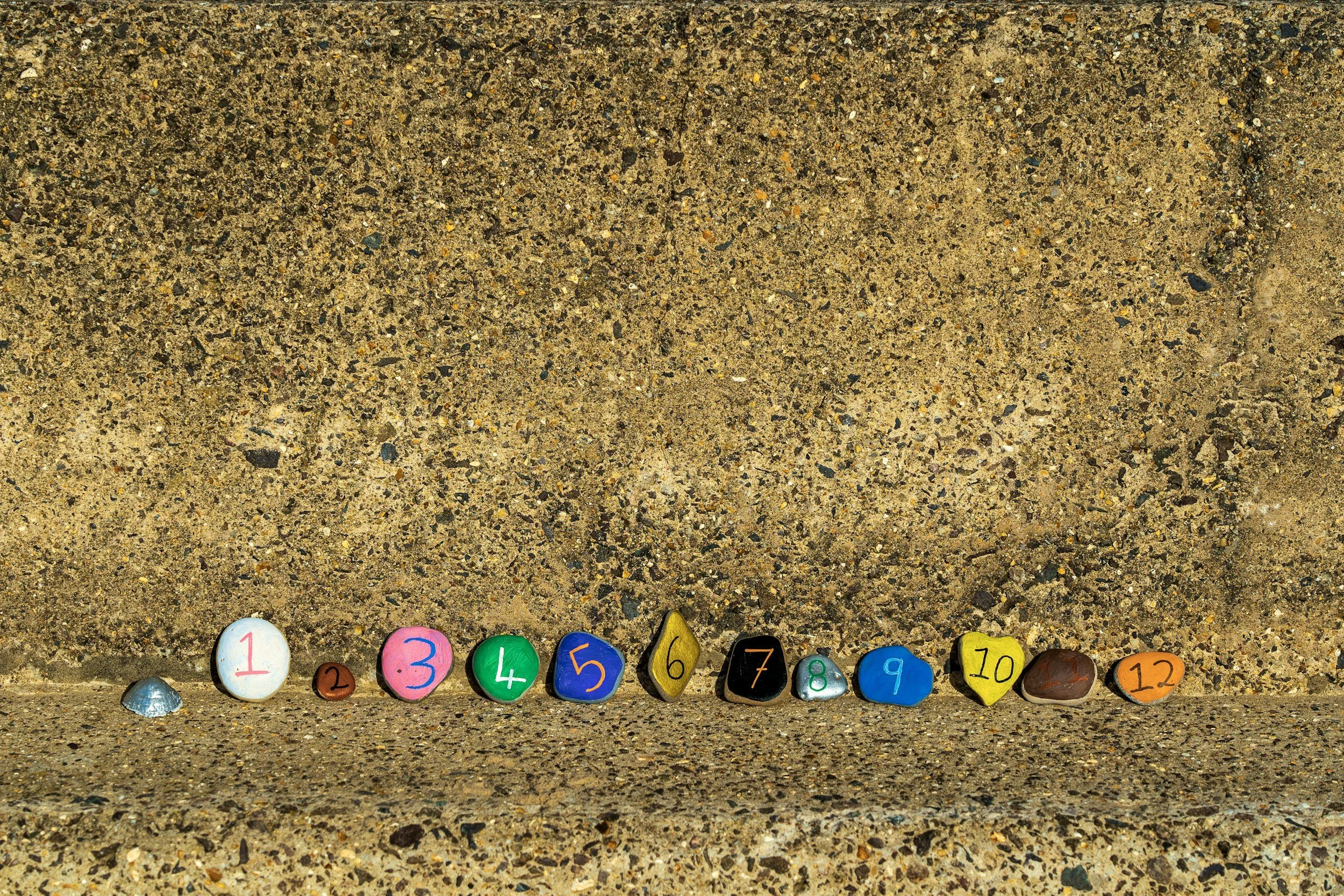 Small painted stones arranged in a line along a concrete surface, numbered 1 through 12 with different colors.