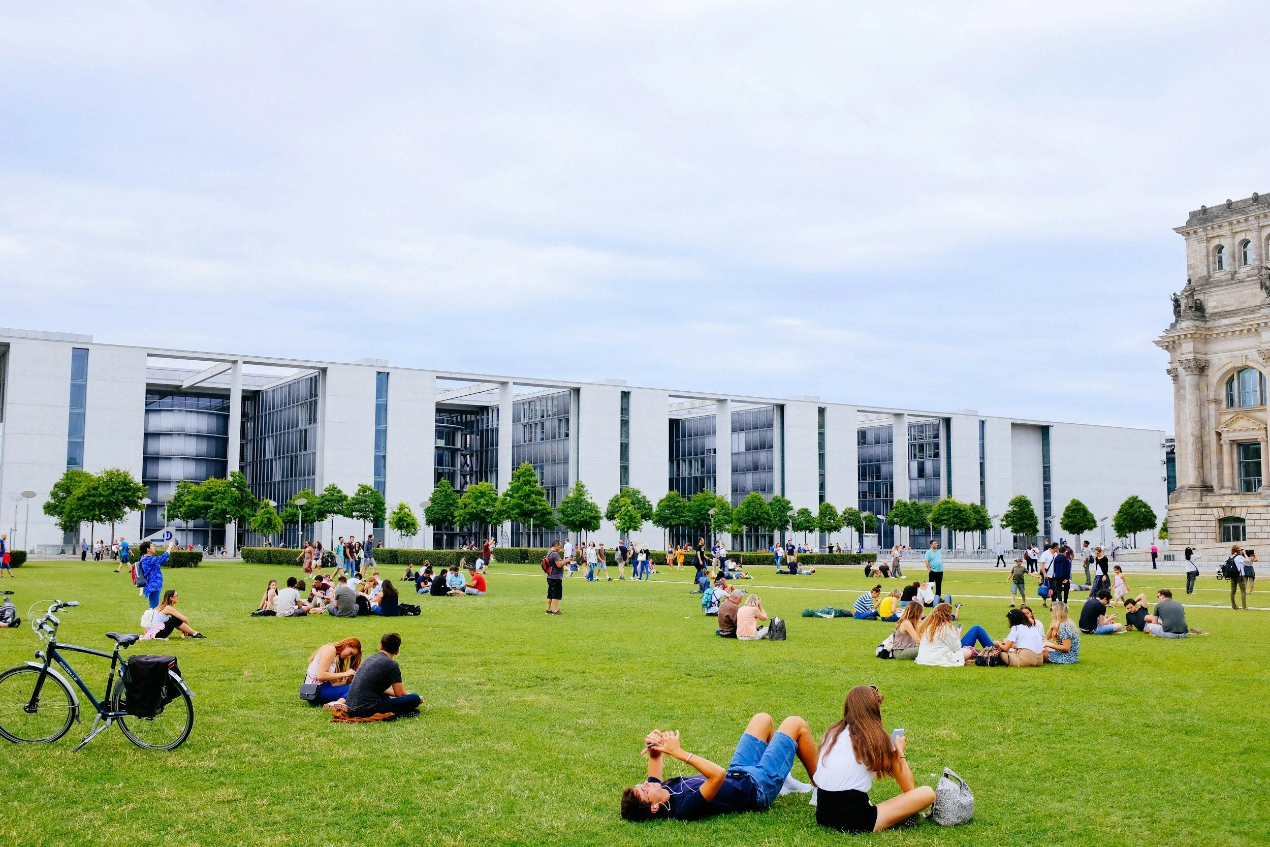 People sitting and relaxing on a grassy field in front of modern office buildings, some using phones, with trees lining the area and a historic building to the right under cloudy skies.
