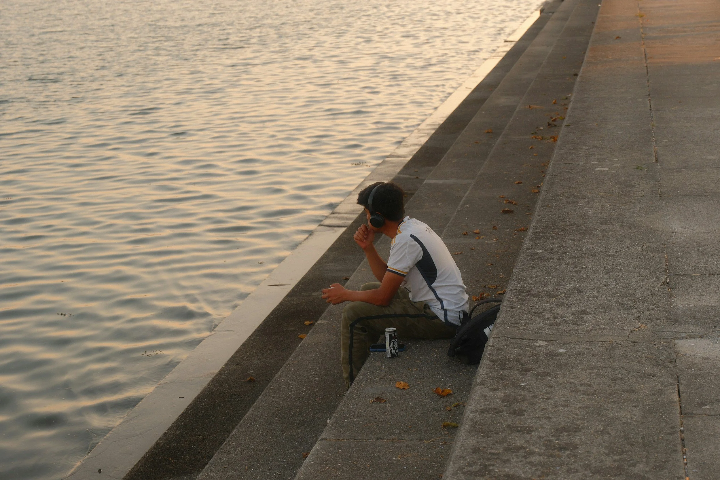 A young man sitting on concrete steps by a body of water, wearing headphones, looking at his phone, with a backpack and a can nearby, during sunset.
