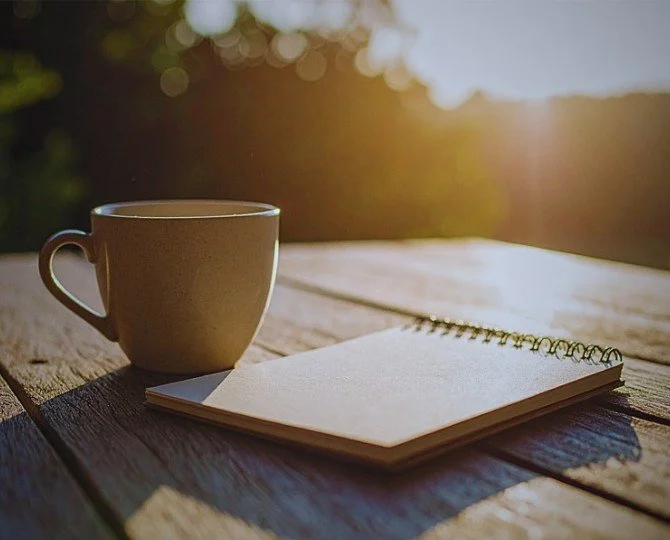 A beige mug and a closed spiral notebook on a rustic wooden table outdoors in sunlight.