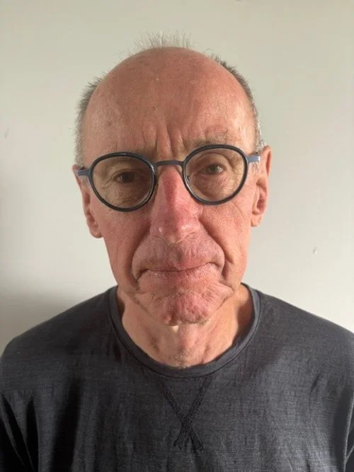 An elderly man with glasses and a serious expression, wearing a dark shirt, standing against a plain light-colored background.