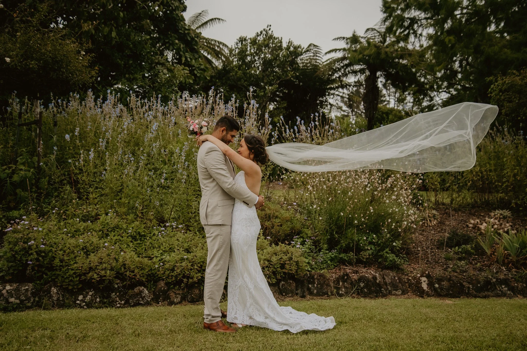 A bride and groom embrace outdoors in a garden, with the bride's veil flowing in the wind and her holding a bouquet of pink and white flowers.