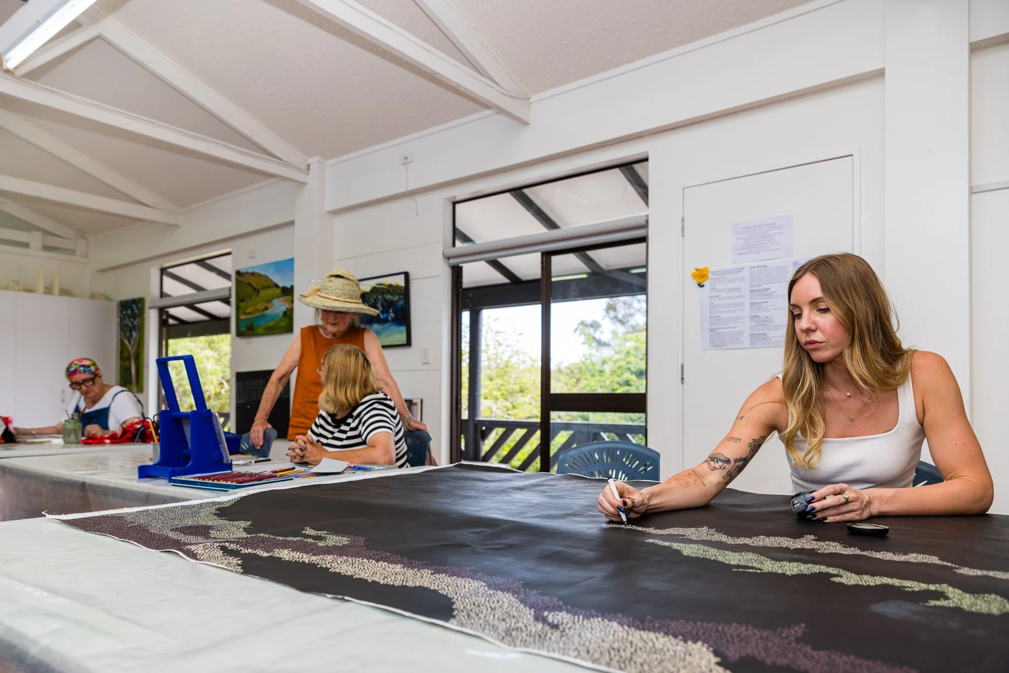 Women working on a large fabric artwork in a bright room with windows and wall art