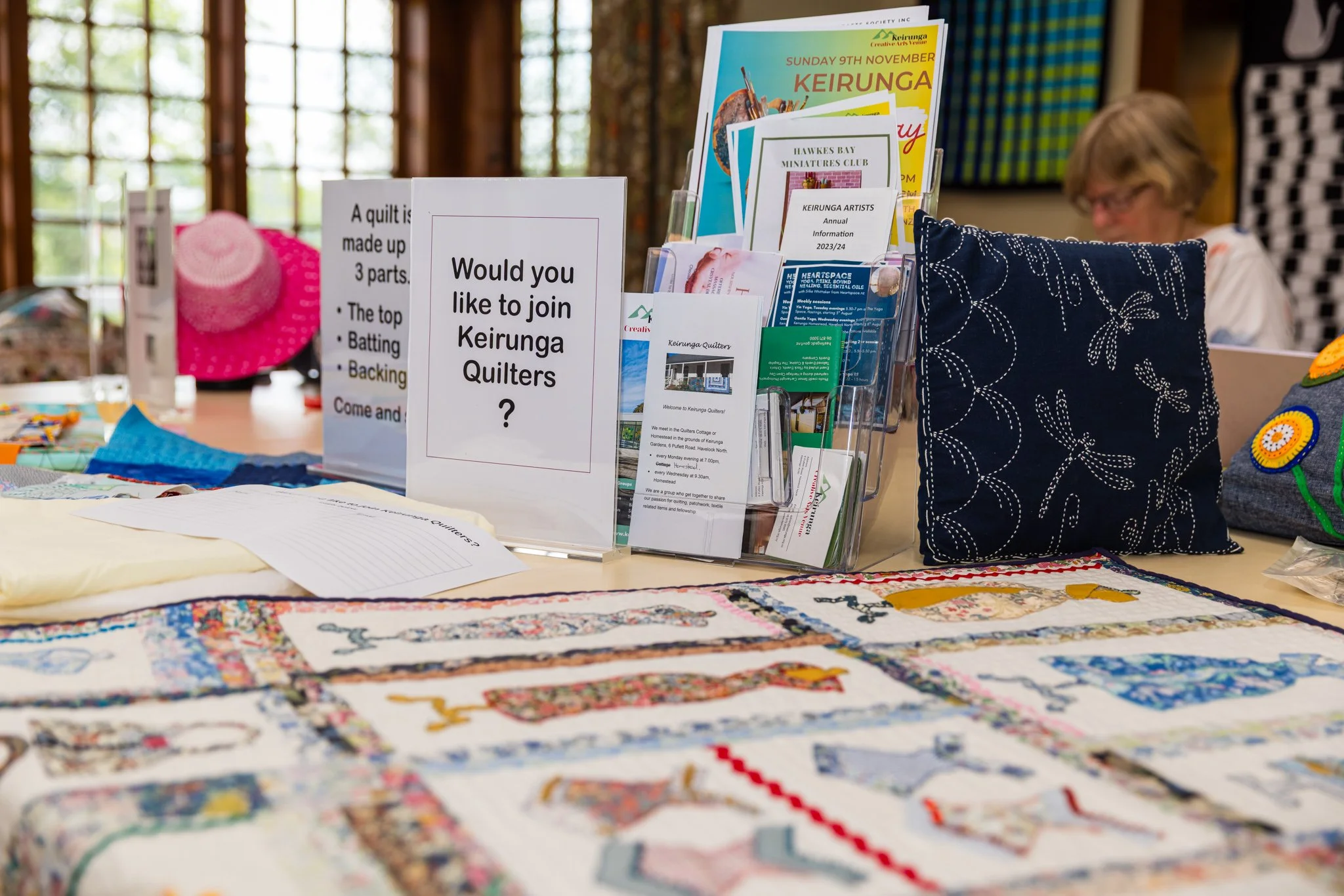 Quilt display table with a sign asking if visitors want to join Keirunga Quilters, along with various brochures and quilted items; background shows a woman and a pink hat.